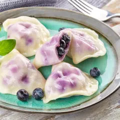 Pyszne pierogi z jagodami, jeden przekrojony, ukazujący soczyste owoce. Podane na turkusowym talerzu z gałązką ziół.