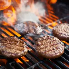 Cztery soczyste burgery z grilla skwierczą na rozgrzanym ruszcie, otoczone płomieniami i dymem.