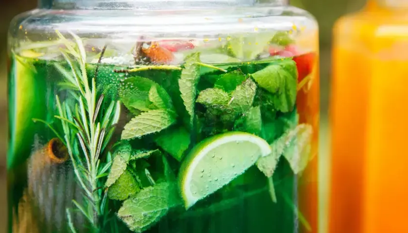 Zielony, orzeźwiający syrop z mięty z dodatkiem limonki i rozmarynu, w dużym słoiku. Obok pomarańczowy napój.