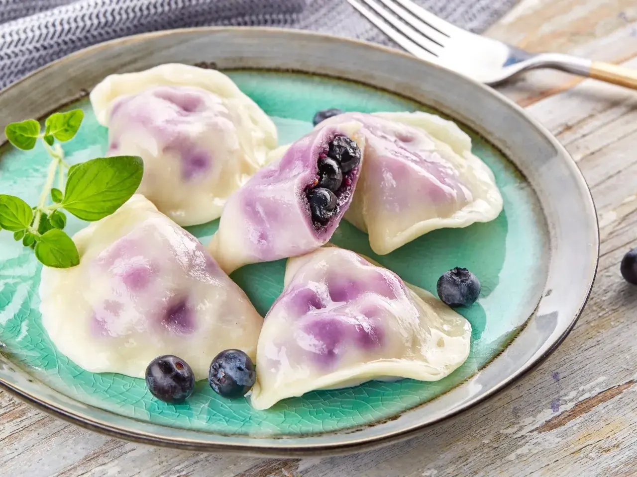 Pyszne pierogi z jagodami, jeden przekrojony, ukazujący soczyste owoce. Podane na turkusowym talerzu z gałązką ziół.