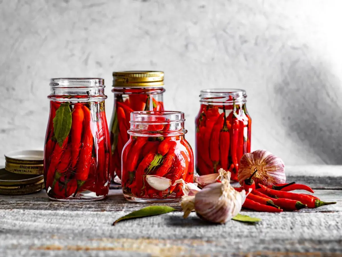 Słoiki z papryczkami chili w zalewie, obok czosnek i świeże papryczki. Idealne do pikantnych potraw.