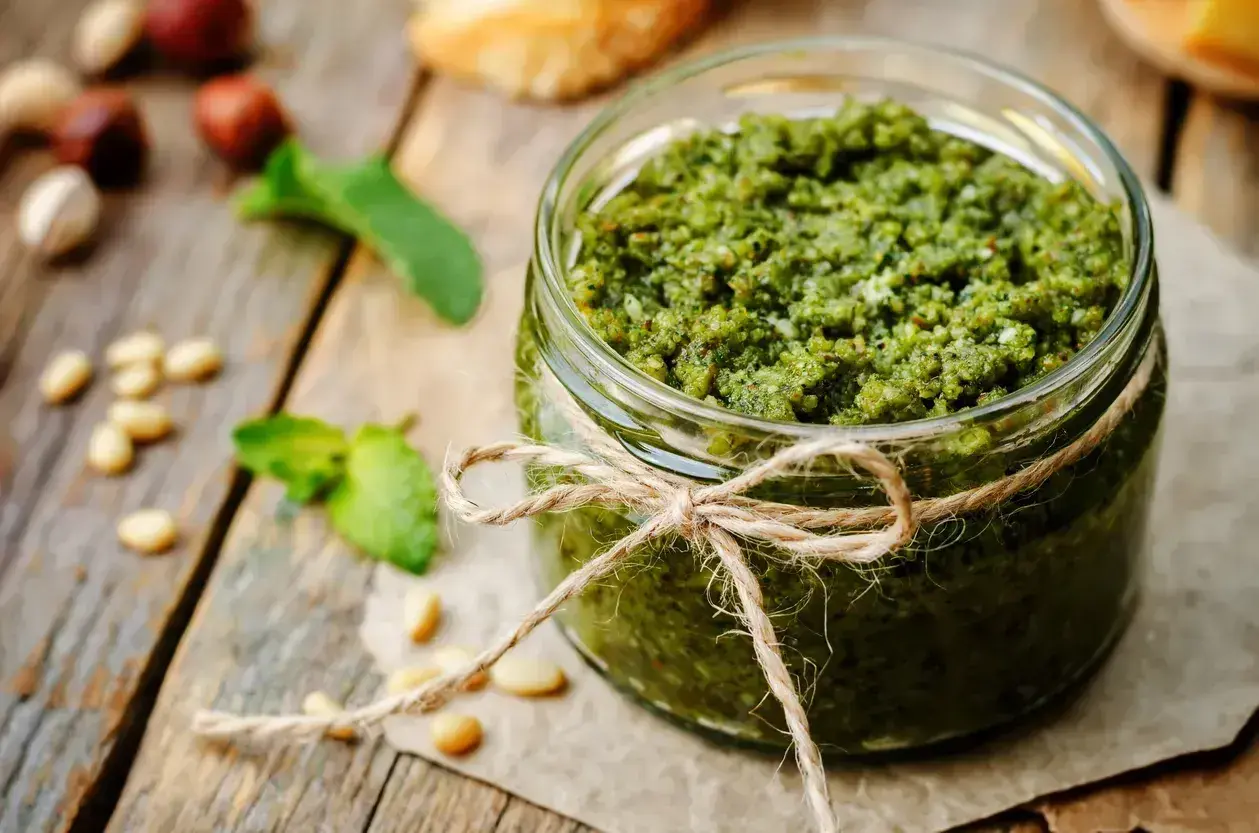 Słoik domowego pesto, ozdobiony sznurkiem, z orzeszkami pinii i listkami bazylii. Idealny przepis, jak zrobić pesto.