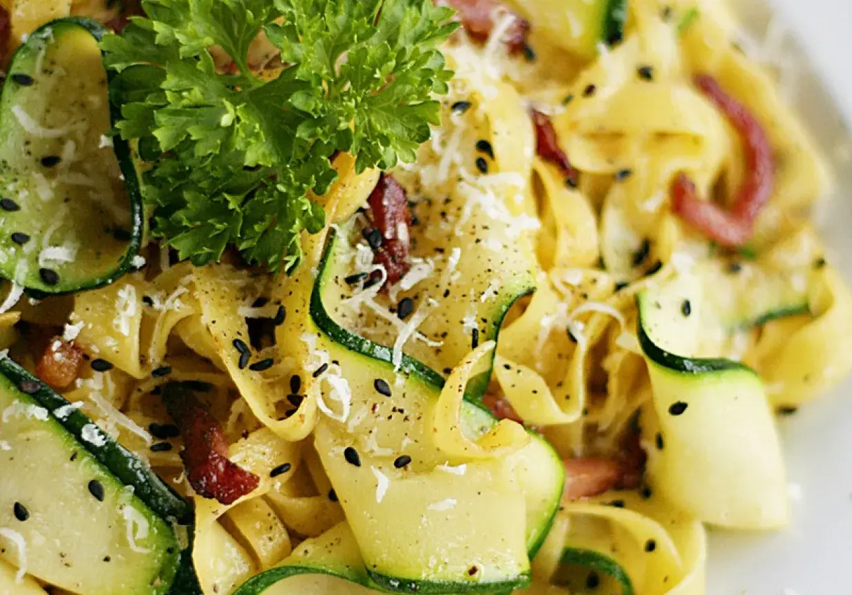 Pyszny makaron z cukinią i boczkiem, posypany parmezanem i czarnym sezamem, udekorowany natką pietruszki.