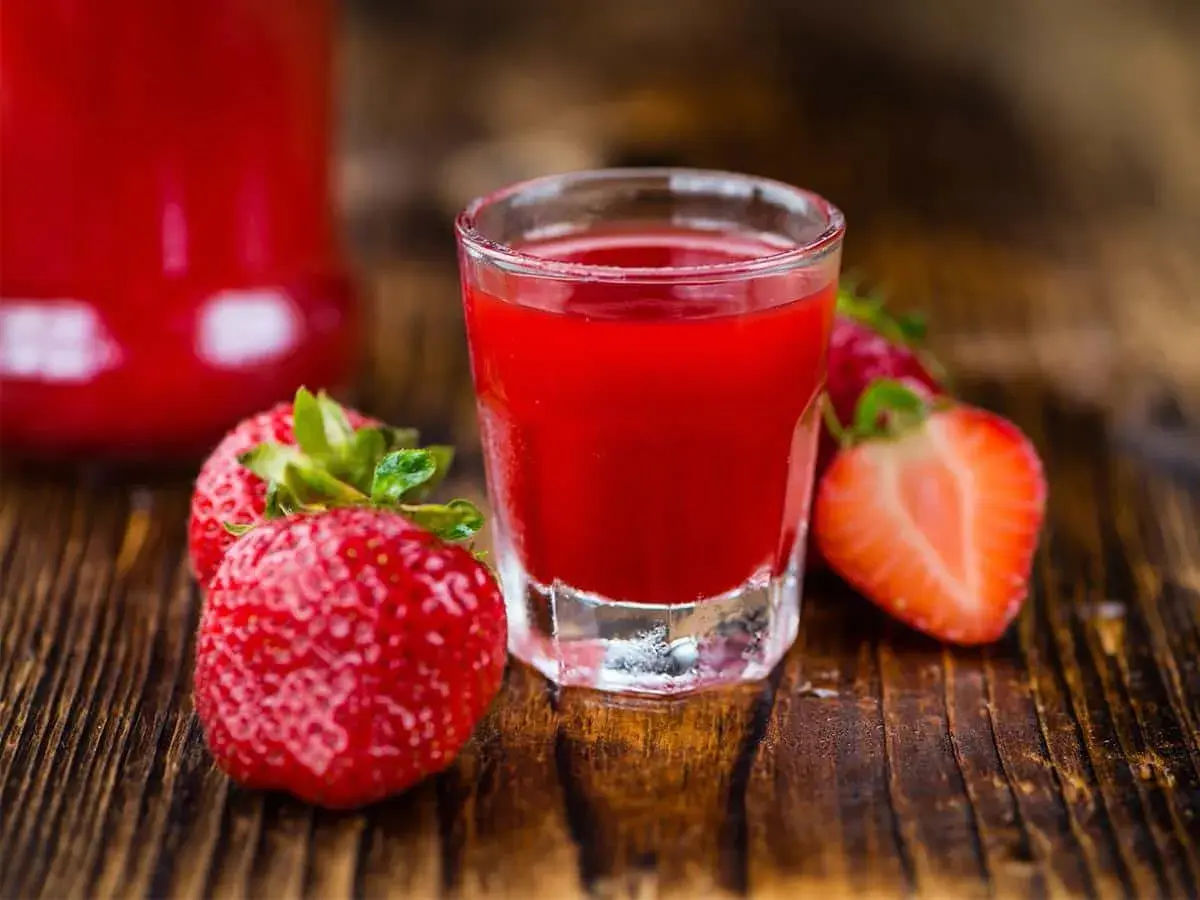 A shot glass filled with vibrant red nalewka z truskawek, surrounded by fresh strawberries on a wooden surface.