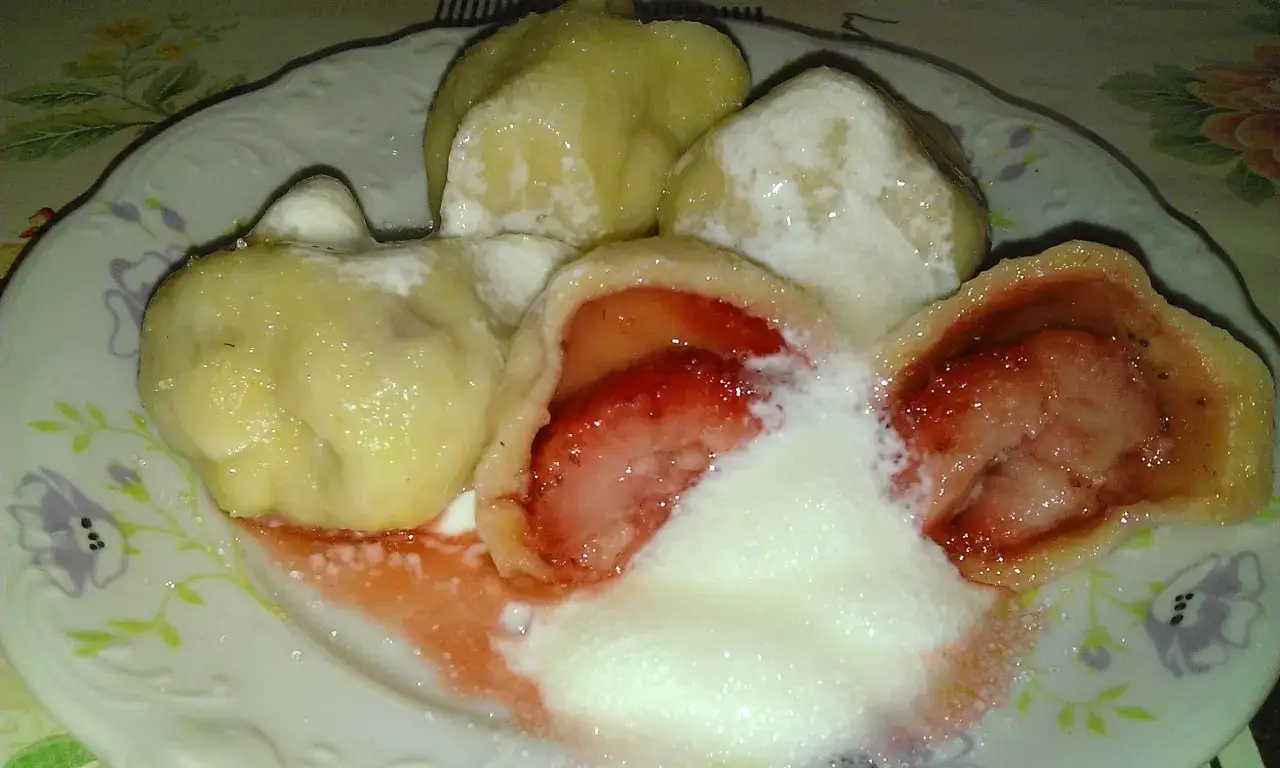 Pyszne pierogi z truskawkami, polane śmietaną i posypane cukrem, na talerzu z kwiatowym wzorem.