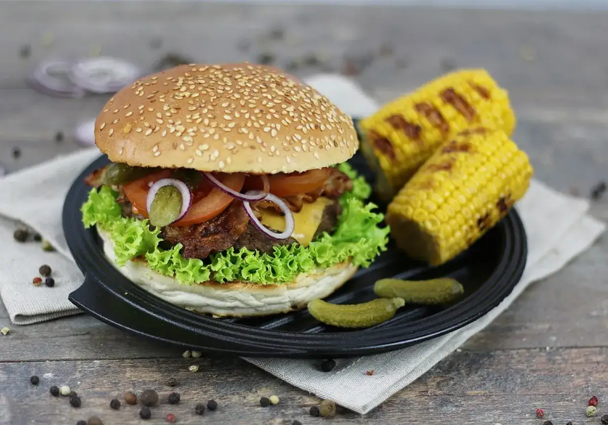 Pyszny burger z grilla z chrupiącą sałatą, pomidorem, serem i bekonem, podany z grillowaną kukurydzą i ogórkami.